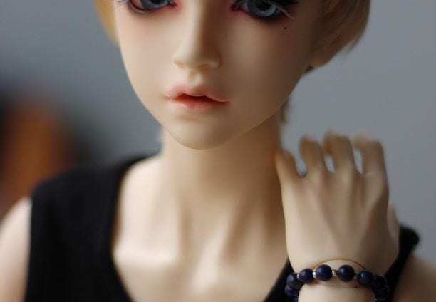 A realistic doll with short blonde hair and striking blue eyes wearing a black sleeveless top. The doll has a neutral expression and is posed with one hand touching the neck, adorned with a black beaded bracelet and a thin brown band.