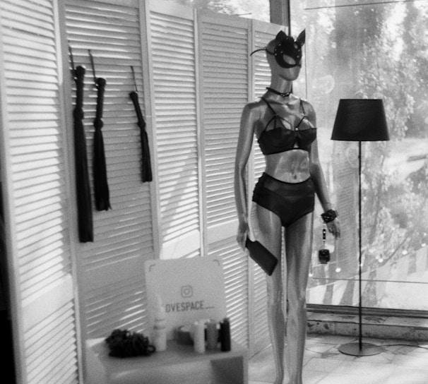 A mannequin dressed in dark lingerie and a mask stands next to a lamp. Nearby, a small table holds various items, including a sign with text. The setting includes paneled dividers and large windows filtering light.